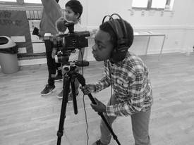 A child stands behind a camera filming something out of the image frame, another child stands nearby to the right